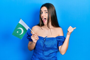 Young brunette teenager holding pakistan flag pointing thumb up to the side smiling happy with open mouth