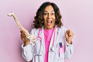 Middle age hispanic doctor woman holding anatomical model of spinal column screaming proud,...