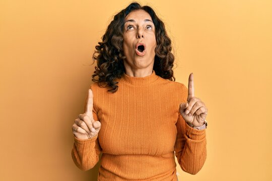 Middle age hispanic woman wearing casual clothes amazed and surprised looking up and pointing with fingers and raised arms.