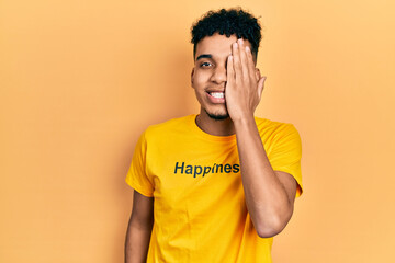 Young african american man wearing t shirt with happiness word message covering one eye with hand, confident smile on face and surprise emotion.