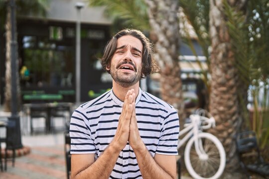 Young hispanic man with beard outdoors at the city begging and praying with hands together with hope expression on face very emotional and worried. begging.
