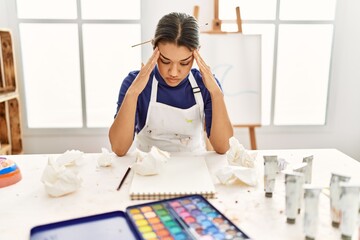 Young latin woman with serious expression sitting on table at art studio