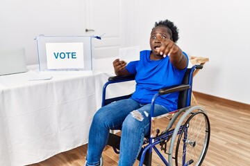 Young african woman sitting on wheelchair voting putting envelop in ballot box pointing with finger...