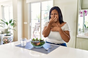 Young hispanic woman eating healthy salad at home smelling something stinky and disgusting,...