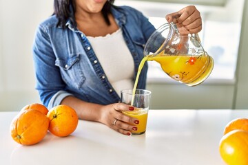 Hispanic brunette woman drinking a glass of fresh orange juice at the kitchen