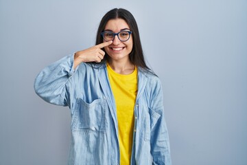 Young hispanic woman standing over blue background pointing with hand finger to face and nose, smiling cheerful. beauty concept