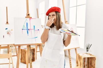 Young artist woman painting on a canvas at art studio smelling something stinky and disgusting, intolerable smell, holding breath with fingers on nose. bad smell