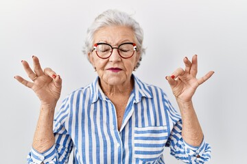 Senior woman with grey hair standing over white background relax and smiling with eyes closed doing meditation gesture with fingers. yoga concept.