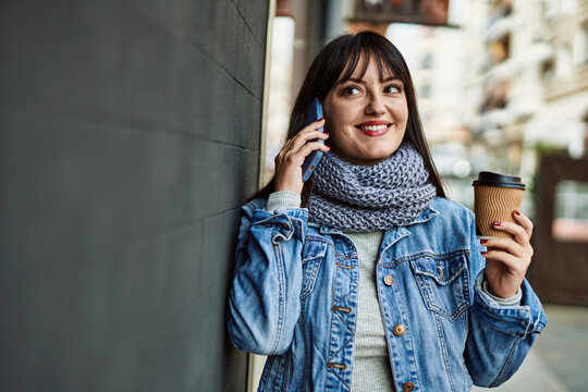 Young brunette woman speaking on the phone and drinking a cup of coffee leaning on the wall