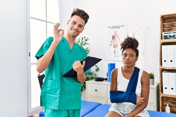 Physiotherapist working with patient wearing arm on sling at the clinic doing ok sign with fingers,...