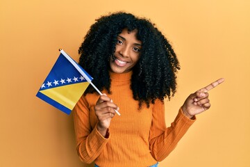 African american woman with afro hair holding bosnia herzegovina flag smiling happy pointing with hand and finger to the side