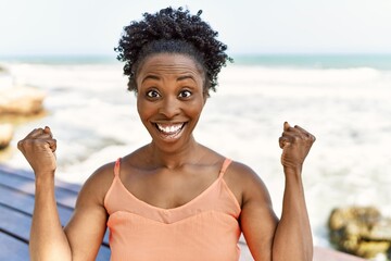 Young african american woman wearing summer clothes standing at the beach screaming proud,...