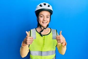 Beautiful brunette little girl wearing bike helmet and reflective vest success sign doing positive gesture with hand, thumbs up smiling and happy. cheerful expression and winner gesture.
