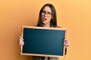 Young hispanic girl holding blackboard in shock face, looking skeptical and sarcastic, surprised with open mouth