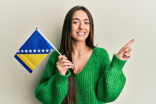 Young hispanic girl holding bosnia herzegovina flag smiling happy pointing with hand and finger to the side