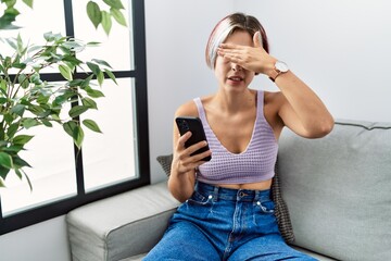 Young beautiful woman using smartphone typing message sitting on the sofa covering eyes with hand, looking serious and sad. sightless, hiding and rejection concept