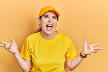 Young hispanic woman wearing delivery uniform and cap crazy and mad shouting and yelling with aggressive expression and arms raised. frustration concept.