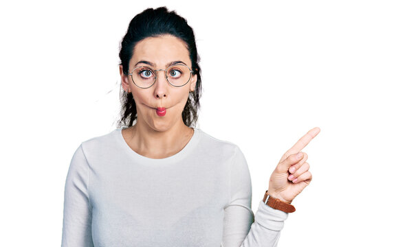 Young hispanic woman pointing with fingers to the side making fish face with mouth and squinting eyes, crazy and comical.