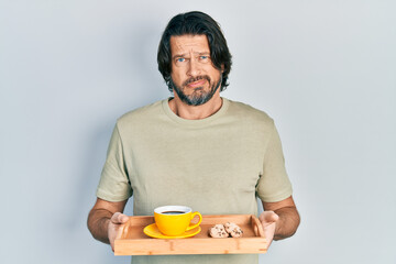 Middle age caucasian man holding breakfast tray with cookies and coffee clueless and confused expression. doubt concept.