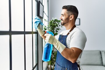 Young hispanic man cleaning window using sprayer at home - obrazy, fototapety, plakaty