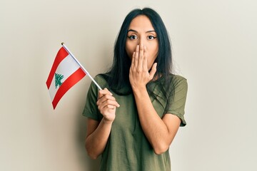 Young hispanic girl holding lebanon flag covering mouth with hand, shocked and afraid for mistake....