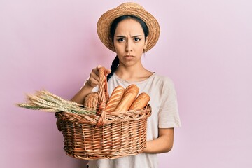 Young hispanic girl holding wicker basket with bread clueless and confused expression. doubt...