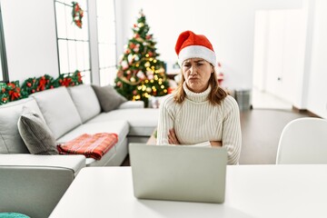 Middle age woman wearing santa claus hat using laptop skeptic and nervous, disapproving expression on face with crossed arms. negative person.