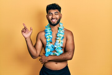 Arab man with beard wearing swimsuit and hawaiian lei smiling happy pointing with hand and finger...