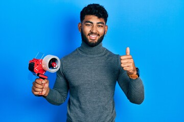 Arab man with beard holding packing tape smiling happy and positive, thumb up doing excellent and approval sign