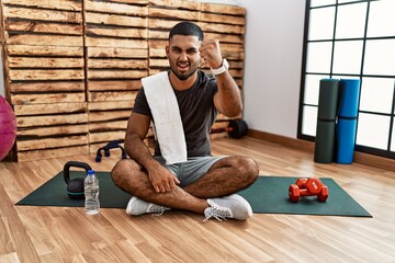 Young indian man sitting on training mat at the gym angry and mad raising fist frustrated and furious while shouting with anger. rage and aggressive concept.