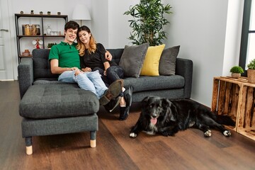 Young caucasian couple smiling happy and hugging sitting on the sofa with dog at home.