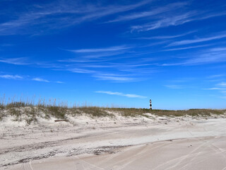 The Cape Lookout, North Carolina lighthouse in the distance from the beach under a blue sky with white clouds