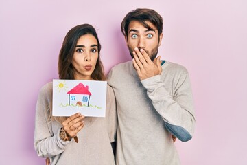 Young hispanic couple holding house draw covering mouth with hand, shocked and afraid for mistake....