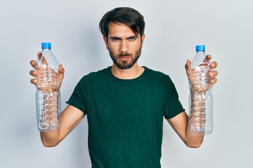 Young hispanic man holding recycling plastic bottles skeptic and nervous, frowning upset because of...