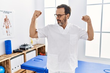 Obraz premium Middle age man with beard working at pain recovery clinic showing arms muscles smiling proud. fitness concept.