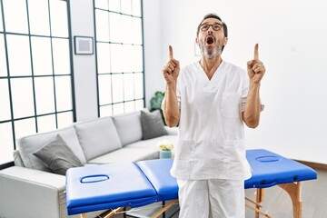 Middle age physiotherapy man working at home amazed and surprised looking up and pointing with fingers and raised arms.