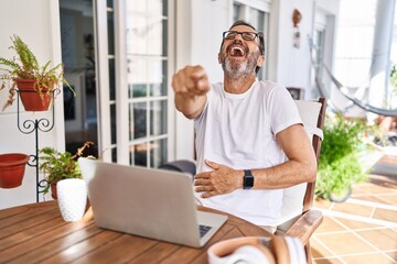 Middle age man using computer laptop at home laughing at you, pointing finger to the camera with...