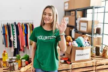 Asian young woman wearing volunteer t shirt at donations stand waiving saying hello happy and...