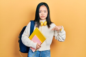 Young chinese girl holding student backpack and books pointing down looking sad and upset,...