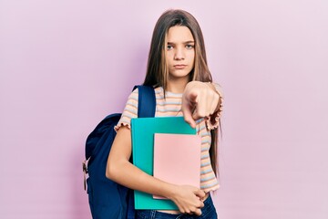 Young brunette girl holding student backpack and books pointing with finger to the camera and to you, confident gesture looking serious