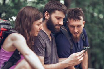 Fototapeta premium Three young friends, two men and a woman, backpacking and going on a hiking trail, looking at their cell phone, navigating through the forest