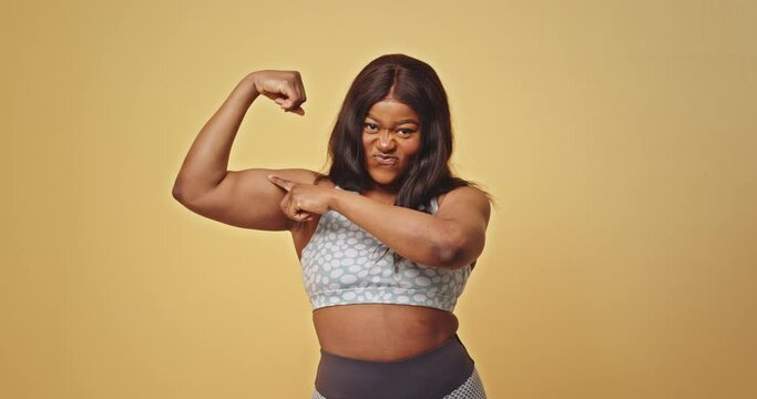 African American sportswoman showing muscles. Plus size black female athlete in activewear smiling and showing biceps after fitness workout against yellow background