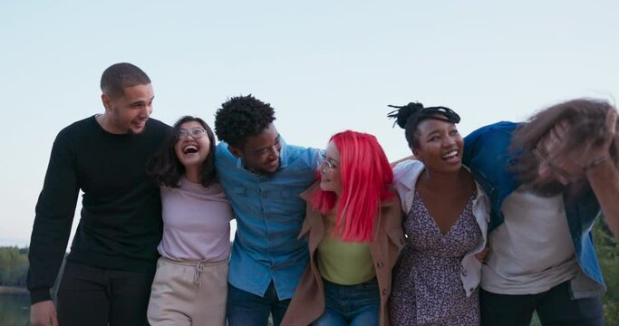 A group of best friends of different nationalities embrace each other in a row, six smiling people lean forward at once looking at the camera, in the background a view of the lake