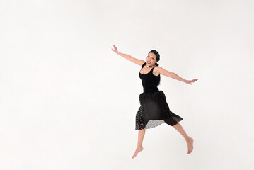 A brunette woman in a black dress barefoot jumped into the air in delight on a white background