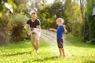 Fototapeta premium Funny little boy with his father playing with garden hose in sunny backyard. Preschooler child having fun with spray of water. Summer outdoors activity for kids.