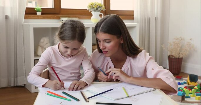 Young loving mother spend leisure with little 6s daughter holding colored pencils drawing together sit table in modern nursery. Teaching and development of children, home hobby, fun, pastime concept