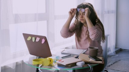 Young woman in glasses working on laptop near window in sunny morning. Lady at messy table with stack of paper and notebooks streatching and massage neck during long sedentary activity