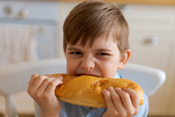 Happy handsome young teenage boy holding and eating freshly baked bread. A hungry boy is biting a big bread, in the kitchen at home. A child with handmade wheat flour bread looks into the camera.