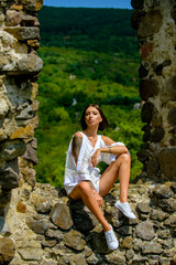 Fashion outdoor photo of gorgeous sensual woman in elegant dress clothes. Woman, fashion model sit on fortress, old castle stones wall.