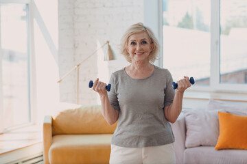 Elderly caucasian old granny aged woman portrait gray haired doing exercises with dumbbells in casual wear at home apartment living room